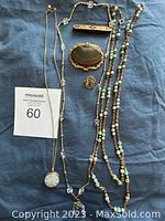 Photo showing the lot of three necklaces and three brooches arranged on blue fabric including the pearl and green bead necklace, crystal pendant necklace, opalescent bead necklace, floral length brooch, agate slice brooch, and rectangular bar brooch.