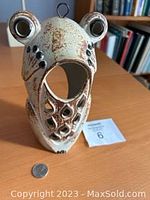 Front perspective of pottery frog candle holder showing large open mouth and eye features, body with decorative cutouts, cream and brown glazing with black accent spots, metal hanging loop on top.