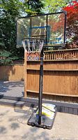 Full view of adjustable basketball hoop with clear backboard, orange rim, and white net set up outdoors on asphalt near a wooden fence.