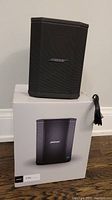 Black Bose S1 Pro speaker resting on its original packaging box with AC power cable coiled next to it