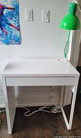White IKEA MICKE desk with green desk lamp attached, photo showing front view with drawers closed and cable cord on floor.