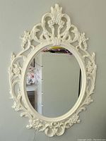 Oval mirror with ornate white flower plastic frame, showing floral and scroll detail. Mirror surface reflects room.