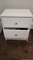 Front view of the IKEA white night table showing two drawers, the top drawer partially open with clothing items inside.