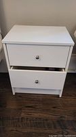 White IKEA night table with two drawers, one drawer slightly open, showing the front, knobs and overall structure on wooden floor.