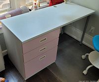White IKEA FRITIDS desk with three pink drawers on a dark wood floor, showing overall desk top and drawer unit.