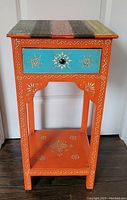 Front view of colorful hand-painted side table with single drawer and decorative motifs on drawer front, legs, and shelf.