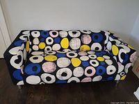 Front view of IKEA Klippan loveseat sofa with black background patterned fabric cover with circles in white, blue, yellow, and pink