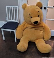 Photo showing large Winnie the Pooh stuffed animal seated on floor next to a white chair for size comparison in a room with dark wood floors.