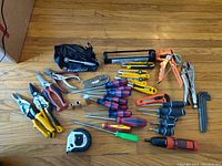 Overview of all tools laid out on wooden floor showing a wide variety of hand tools including screwdrivers, pliers, socket sets, caulking gun, measuring tape and others