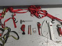 Photo of two red ratchet straps with hooks, plumbing tools including pipe wrenches and pliers, a metal caliper, a small digital device, a white power strip, and adjustable wrench lying on a gray surface.