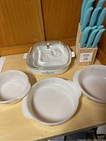 Photo showing three Corning Ware bakeware pieces: round dish, large bowl, smaller bowl, along with square casserole dish with lid and blue-handled knife set in wooden block.
