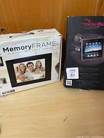 Photo showing the boxed MemoryFrame digital photo frame and Rocketfish iPad 2 car mount side by side on a wooden surface.