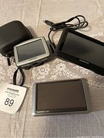 Three GPS units on a grey patterned tablecloth showing TomTom and Garmin brands, with charging cables and carrying case.