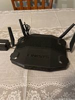 Black Linksys WRT32X wireless router with four antennas on gray lace tablecloth, accompanied by power adapter.