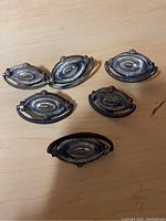 Five vintage oval metal drawer handles arranged flat, showing ornamental details and darkened patina.
