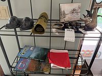 Wide view showing beaver figurines, driftwood carving, pins, framed painting, books, flag, and woven mat on black shelving unit.