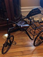 Side view of black metal tricycle wine rack on wood floor showing seat, handlebars, and metal basket space for bottles