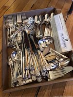 Box filled with assorted silver plated flatware including forks, knives, spoons and serving pieces all with ornate floral pattern on handles.