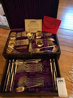 Full view of two-tiered black fitted canteen containing various pieces of silver plated flatware including forks, spoons, knives, and serving utensils. Purple fabric lining inside box, and red pouch alongside.