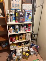 White pressboard shelving unit filled with a variety of household cleaning products including spray cans, liquid bottles, and boxes.