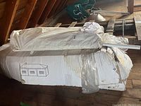 Packed large backyard party tent stored atop old pallets in attic space wrapped in plastic and cardboard packaging showing line drawing of tent.