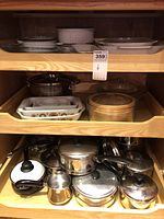 Wooden cupboard shelves showing various CorningWare French white casserole dishes and bowls with lids on the upper shelves, clear glass steamer tray, and a variety of stainless steel pots and pans with black handles and clear glass lids on lower shelves.