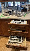 Overview of kitchen counter showing mugs, kettle, espresso machine, creamers, and espresso tea set, as well as three open drawers containing kitchen utensils and flatware.