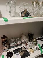 Wide view of lot on white shelves showing ceramic stein, house cookie jar, porcelain pitcher, shakers, and decorative items