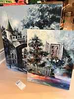 Two original paintings visible, one of a castle or stone structure with a tower, and one of a house with open shutters and white picket fence.