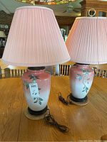 Two matching table lamps showing ceramic bases with gradient dusty rose to white finish, hand-painted bird and leaf appliqué details and dusty rose pleated shades on a wooden table.