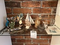 Full view of assorted souvenir collectibles including birds, sailboats, blown glass horse, glass jar, and metal/bone figurines on glass shelf against brick wall.