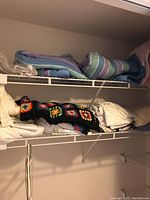 Wide view of shelf containing linens including folded striped comforter, white sheets, and black crocheted afghan with colorful squares.