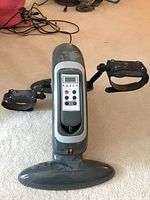 Top and front view of the grey Ivation electric mini exercise bike on carpet, showing black adjustable pedals with straps and control panel on top.