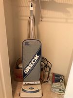 Photo of Oreck upright vacuum with blue and beige bag covering and power cord wrapped on the back. Vacuum is standing upright on carpet near door.