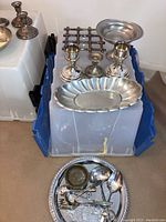 Silver plated items including candle sticks, trivet, small candle holder, serving platter, candy dishes, and small serving pieces laid on a plastic container.