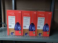 Three boxed Tiella Fiero Pendant Packs on a metal shelf, each box showing an image of the pendant light with a blue glass shade