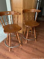 Two wooden counter height swivel chairs with spindle backs, curved top rail, and metal circular footrests, displayed on hardwood floor near kitchen cabinetry.