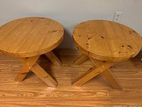 Two handmade round wooden side tables with round tops and crossed legs, placed adjacent on wooden floor.