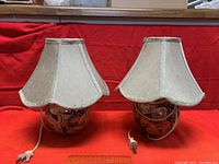 Two small ceramic lamps with beige fabric shades placed side by side against a red fabric background.