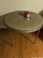 Full view of vintage round folding card table with beige textured laminate top, showing metal legs and wood floor beneath. Candy tin placed on table top for scale and decoration.