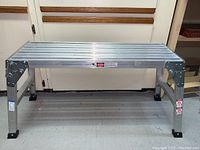 Front view of the aluminum platform showing rectangular slatted standing surface and foldable legs with rubber feet.
