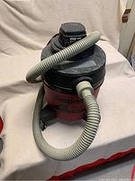 Front perspective of red and black Shop Vac with attached gray hose, showing the unit's upper, middle, and lower parts including lid and caster wheels.