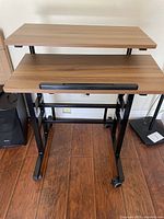 Photo taken showing a two-tier compact computer desk with a light wood surface and black metal frame with caster wheels.