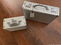 Two closed silver hard side cases with combination locks on a wooden floor.