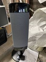 Tall rectangular Lasko ceramic pedestal heater with black and gray front grille, standing upright on a circular base. The remote is visible lying at the base along with instruction manual.