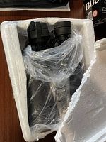 Binoculars inside a custom styrofoam box wrapped in clear plastic, front view showing eyepieces and strap.