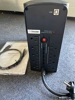 UPS shown from rear side featuring 9 outlets, a USB port for communication, and a red resettable circuit breaker button. Power cord attached and Amazon Basics manual plus USB cable packaged separately nearby.