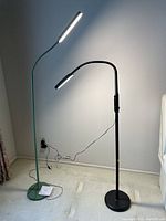 Two LED floor lamps lit, one in green and one in black, showing their design with flexible necks and light bars.
