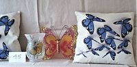 Photo showing two butterfly print cushions, a metal butterfly wall art, and a glass candle holder in front of a beige backdrop.