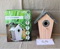 Front view of bird house with tin roof and circular entry hole, next to original packaging, showing branding and design.